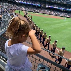 Heartfelt Tribute: Young Girl's Salute to Servicemen Captivates Crowd at Baseball Game